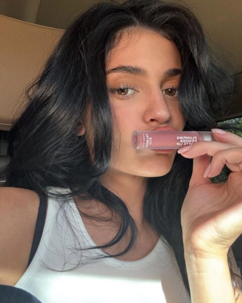 Zdjęcie celebrytki Kylie Jenner. Siedzi w samochodzie, ubrana jest w biały top bez rękawów. Ma ciemne włosy rozpuszczone na ramiona. Przykłada do ust swoją pomadkę do ust.