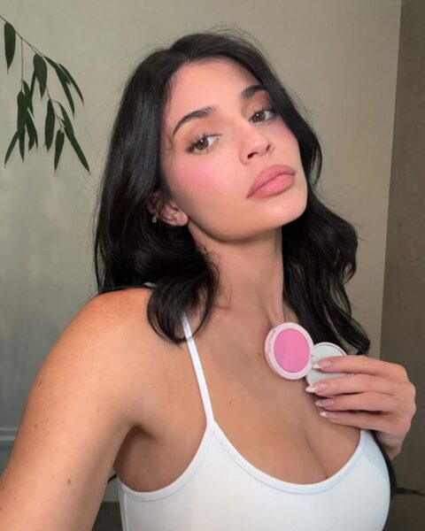 Zdjęcie celebrytki Kylie Jenner, która zrobiła sobie selfie, by promować róże jej własnej marki Kylie Cosmetics. Ma mocny makijaż z wyrazistym różem i efektownie pomalowane usta oraz sztuczne rzęsy. Włosy ciemne, rozpuszczone. Trzyma w ręce róż i patrzy w obiektyw. Ubrana jest w biały top na ramiączkach.