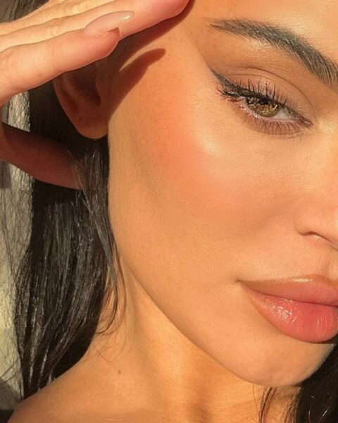 Zdjęcie celebrytki Kylie Jenner z mocnym zbliżeniem na jej twarz. Przykłada rękę do skroni i lekko unosi kącik wymalowanego i wytuszowanego oka. Widać też makijaż ust Kylie Jenner z brązową konturówką. Widać też naturalną strukturę jej skóry.