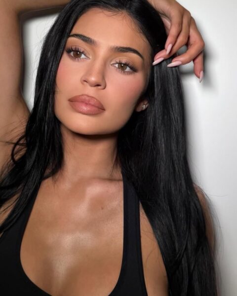 Zdjęcie celebrytki Kylie Jenner. Jest ubrana w czarny top na ramiączkach. Ma ciemne, proste włosy rozpuszczone na ramiona. Opiera rękę o głowę. Na twarzy nosi wyrazisty makijaż z podkreślonymi ustami.