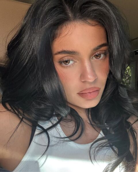 Zdjęcie celebrytki Kylie Jenner, która siedzi w samochodzie i zrobiła sobie selfie. Ma ciemne włosy rozpuszczone na ramiona. Jest ubrana w biały top bez rękawów. Ma delikatny makijaż z podkreślonymi ustami.