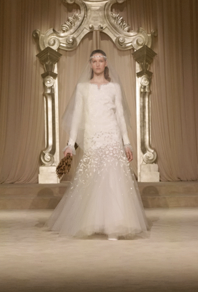 Suknia ślubna CHANEL z kolekcji Haute Couture Jesień-Zima 2025/26 – długa, tiulowa kreacja z haftem przypominającym kłosy pszenicy, zestawiona z welonem i symbolicznym bukietem z suszonych zbóż; modelka prezentuje look w scenerii złocistej, teatralnej oprawy.