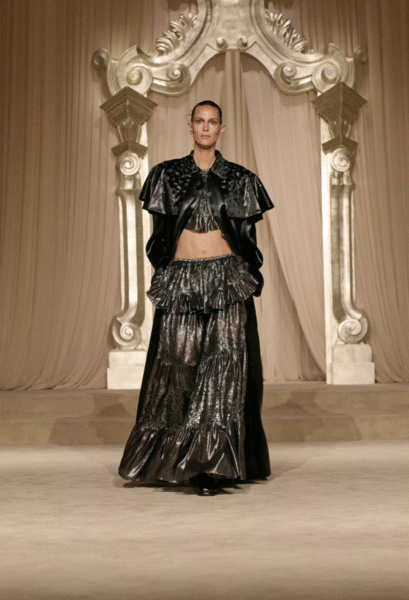 Czarny, metalicznie połyskujący look z kolekcji CHANEL Haute Couture FW25/26 – długa, falbaniasta spódnica i krótki top z peleryną w stylu bolerka, ozdobioną perełkami i haftami, kontrastują z klasyczną architekturą w tle; stylizacja balansująca między gotykiem a romantyzmem.