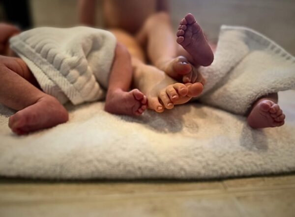 Zbliżenie na stópki trójki małych dzieci leżących na miękkim ręczniku. Dwoje z nich ma nogi przykryte białymi ręcznikami, a jedno leży na środku z widocznymi palcami stóp. Zdjęcie ma miękki, ciepły klimat.