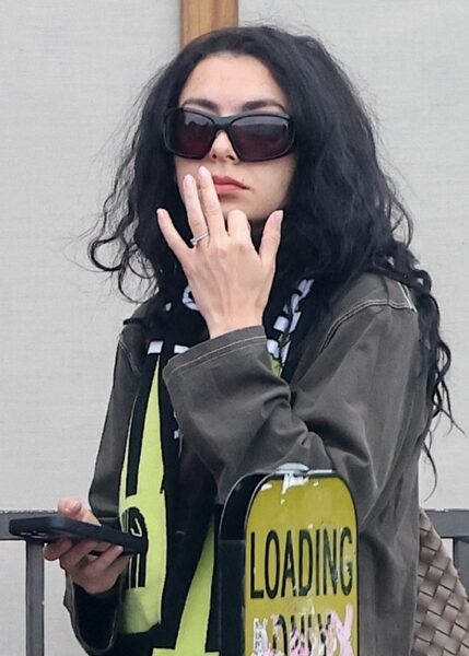 Zbliżenie na Charli XCX, która unosi rękę z widocznym pierścionkiem zaręczynowym, trzymając jednocześnie telefon. Ubrana jest w ciemną kurtkę w stylu militarnym oraz szalik z graficznym motywem w jaskrawych kolorach. Jej twarz zasłaniają duże, zakrywające pół twarzy okulary przeciwsłoneczne. Charli wygląda nonszalancko i tajemniczo – jej styl to połączenie estetyki cyber-grunge z wpływami popkultury lat 2000. Zdjęcie uchwycone w przestrzeni miejskiej, prawdopodobnie w trakcie codziennych obowiązków lub przed wejściem do samochodu.