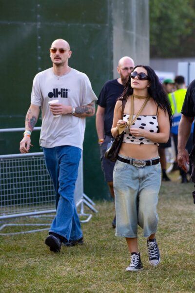 Charli XCX i George Daniel na terenie festiwalu muzycznego, prawdopodobnie w trakcie wydarzenia typu Glastonbury lub Lollapalooza. Charli ma na sobie krótką, asymetryczną bluzkę w czarne grochy, niskie, szerokie jeansowe spodenki typu bermudy, pasek z ozdobną klamrą oraz klasyczne czarne trampki Converse. Jej styl wpisuje się w festiwalową estetykę DIY z lat 2000. George ma na sobie jasną koszulkę z nadrukiem "DHLiii" oraz luźne niebieskie jeansy, stylizowane na lata 90. Para wygląda na zrelaksowaną i zgrzaną z klimatem wydarzenia.