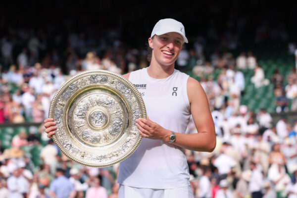 Iga Świątek z uśmiechem pozuje z trofeum Wimbledonu 2025 na tle trybun kortu centralnego. Jej stylowy zegarek Rolex i sportowy strój On Running uzupełniają zwycięski look liderki WTA.