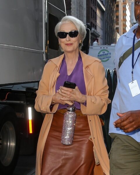 Meryl Streep jako Miranda Priestly: Meryl Streep w roli Mirandy Priestly na planie filmu „The Devil Wears Prada 2” w Nowym Jorku; ubrana w długi karmelowy trencz, fioletową bluzkę z dekoltem w szpic oraz skórzaną ołówkową spódnicę w kolorze koniakowym. Trzyma w dłoniach telefon i błyszczącą, kryształową butelkę, ma na sobie ciemne okulary przeciwsłoneczne i złotą biżuterię.