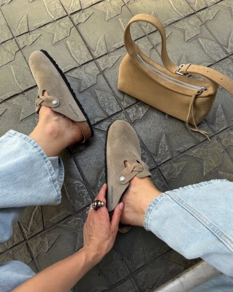 Zbliżenie na osobę noszącą beżowe klapki Birkenstock z zamszu, siedzącą na chodniku w jasnych jeansach. Obok leży mała, beżowa torebka z suwakiem. Jedna dłoń trzyma but, widoczny także ozdobny pierścionek.