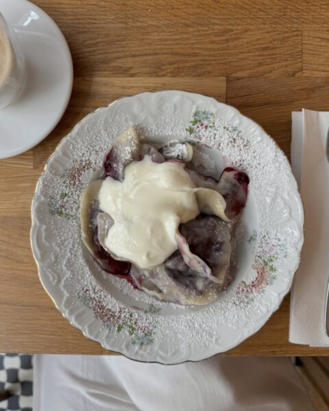 Talerz z pierogami z jagodami, polanymi śmietaną i posypanymi cukrem pudrem, podany na ozdobnym porcelanowym talerzu, obok filiżanki kawy i sztućców na drewnianym stole.