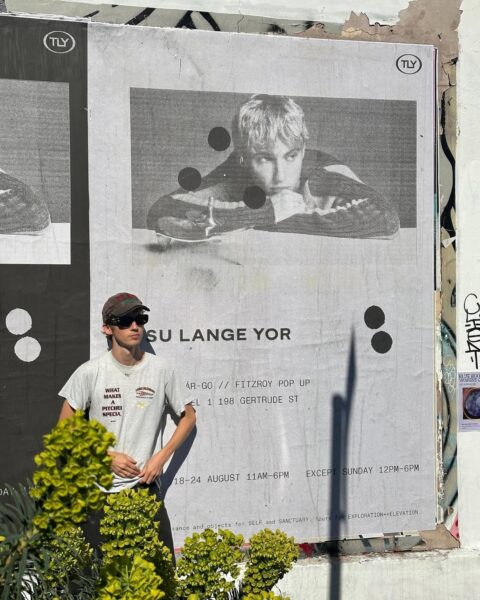 Duży plakat promujący markę Tsu Lange Yor rozwieszony na miejskiej ścianie z graffiti. Na plakacie czarno-białe zdjęcie Troye’a Sivana w tej samej pozie co na zdjęciu portretowym – oparty na stole, patrzy zamyślony. Obok niego młody mężczyzna w okularach przeciwsłonecznych i czapce z daszkiem, pozujący przy plakacie. Widoczna lokalizacja pop-upu w Fitzroy oraz daty wydarzenia.