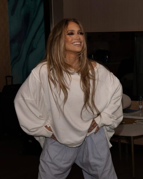 Jennifer Lopez pozuje w garderobie w luźnej, kremowej bluzie i jasnoniebieskich, szerokich spodniach z wysokim stanem. Ma rozpuszczone, starannie ułożone włosy i promienny uśmiech na twarzy, co nadaje zdjęciu ciepłego, naturalnego charakteru. W tle widać fragment hotelowego wnętrza — designerską ścianę, stół z krzesłem i butelkę wody, co sugeruje moment relaksu za kulisami tuż przed lub po występie.