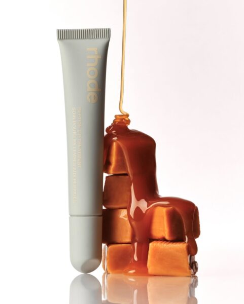 Błyszczyk do ust rhode – Peptide Lip Treatment – w odcieniu Salted Caramel, ustawiony obok kostek karmelu, po których spływa złocisty, gęsty sos karmelowy.
