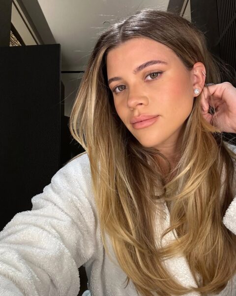 Kobieta w białym szlafroku robi selfie w dobrze oświetlonym wnętrzu; ma długie, rozpuszczone blond włosy, delikatny makijaż i nosi kolczyki z cyrkoniami.