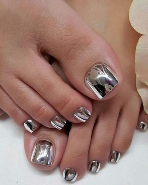Zdjęcie stóp z pedicure w kolorze srebrnym, metalicznym wykończeniu. Stopy pozują ułożone jedna na drugiej tak, by prezentować pedicure.