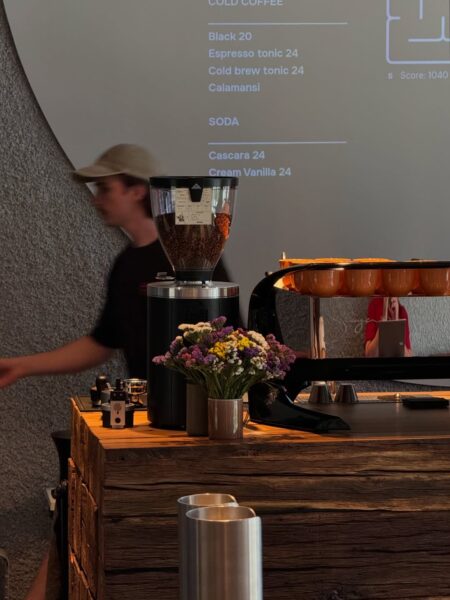 Wnętrze kawiarni z drewnianym barem, na którym stoją młynek do kawy z ziarnami, bukiet kolorowych kwiatów i ekspres z rzędem pomarańczowych filiżanek. W tle widać rozmytą sylwetkę baristy w czapce oraz fragment menu wyświetlonego na ścianie.