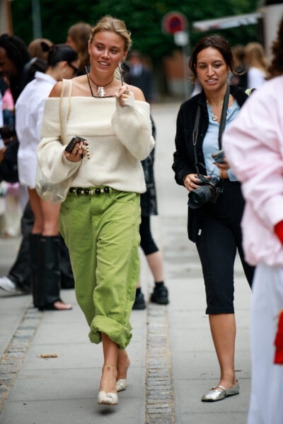 Street style w codziennym wydaniu – kobieta w luźnym, kremowym swetrze odsłaniającym ramiona, jasnozielonych spodniach z wywiniętymi nogawkami, w beżowych balerinach i z czarnym paskiem z ozdobnymi kulkami, idąca w towarzystwie innej osoby z aparatem.