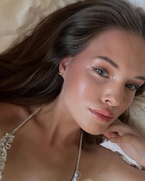 Zdjęcie kobiety, która ma gładką, lśniącą twarz po zabiegach kosmetycznych. Ma ciemne włosy. Leży na łóżku i robi sobie selfie.
