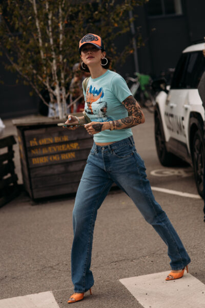 Jeansy na jesień 2025: Street style Kopenhaga 2026 – kobieta w jeansach z prostą nogawką w odcieniu vintage blue, turkusowym T-shircie z nadrukiem i pomarańczowej czapce z daszkiem. Stylizacja dopełniona pomarańczowymi szpilkami i złotą biżuterią, uchwycona na ulicy podczas fashion week.