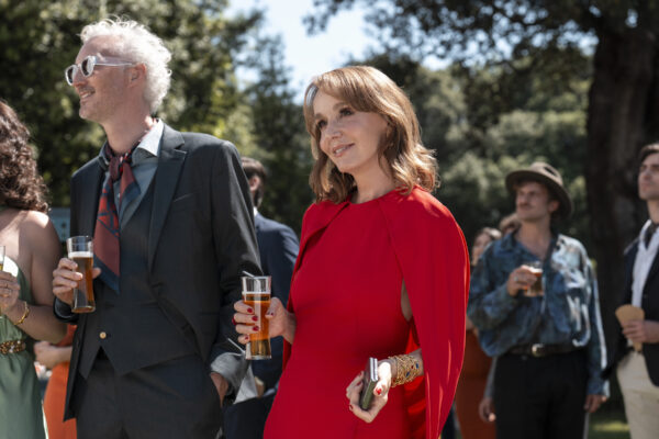 Postać ubrana w efektowną czerwoną kreację z peleryną trzyma drinka i uśmiecha się w stronę rozmówców, stojąc w ogrodzie podczas eleganckiego przyjęcia w plenerze.