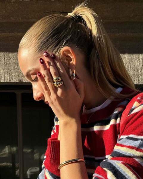 Zdjęcie kobiety stojącej bokiem. Ma blond włosy związane w wysoki kucyk. To fryzura liftingująca. Ubrana jest w czerwony sweter w paski, dużo pierścionków na palcach oraz czerwone paznokcie. Przykłada rękę do twarzy, ale patrzy gdzieś w dół.