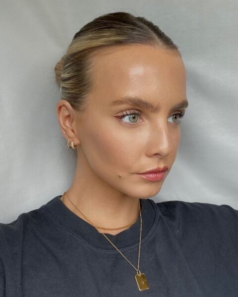 Zdjęcie kobiety w blond włosach gładko związanych w kok. Patrzy w bok. Ubrana jest w czarny T-shirt. Na szyi ma złoty naszyjnik, a w uszach złote kolczyki.
