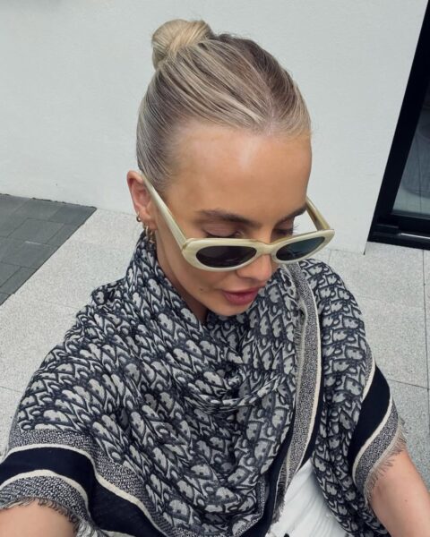 Zdjęcie kobiety, która zrobiła sobie selfie od góry. Ma blond włosy związane gładko w kok, na nosie beżowe okulary przeciwsłoneczne, a na ramionach czarno-białą chustę. Ubrana jest w białą kamizelkę z czarnymi guzikami.