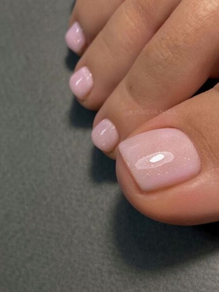 Zbliżenie na starannie wykonany pedicure w odcieniu pudrowego różu z delikatnym, perłowym połyskiem. Paznokcie u stóp mają krótki, kwadratowy kształt i subtelny, elegancki połysk, który podkreśla zadbaną skórę i klasyczną estetykę stylizacji. Pedicure emanuje minimalizmem i luksusem w wersji soft.