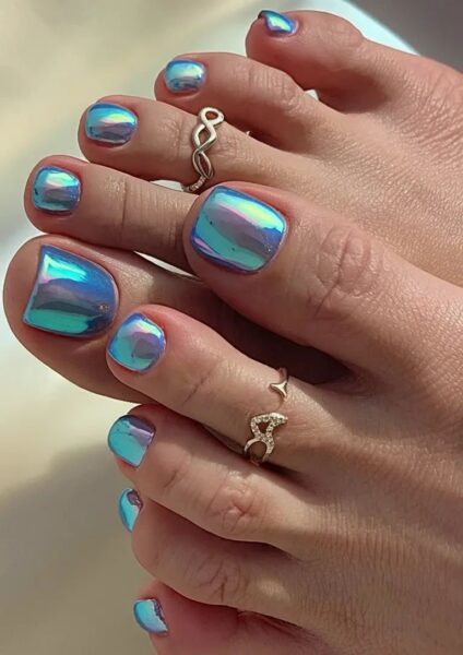 Pedicure w stylu „mermaid chrome” – paznokcie u stóp pokryte są opalizującym lakierem w odcieniach niebieskiego, zieleni i różu z efektem tafli lustra. Stylizacja przyciąga wzrok intensywnym błyskiem i futurystycznym wykończeniem. Uwagę przyciągają również złote pierścionki na palcach, które dopełniają morski klimat.