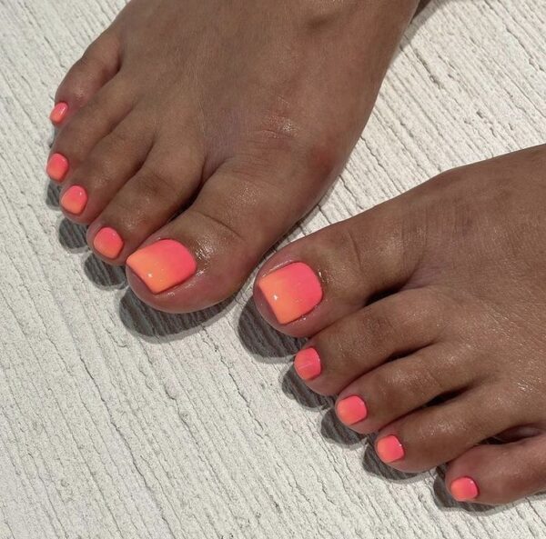 Letni pedicure w intensywnym neonowym odcieniu pomarańczowo-różowego gradientu. Stylizacja inspirowana energetycznym zachodem słońca, widoczna na tle jasnego, teksturowanego podłoża. Kolory emanują ciepłem i radością, przyciągając uwagę żywą, soczystą barwą idealną na lato.