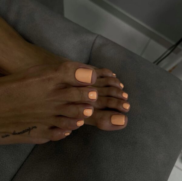 Pedicure w neutralnym, brzoskwiniowym odcieniu na ciemniejszej karnacji, wykonany na krótkich, kwadratowych paznokciach. Stylizacja prezentuje się elegancko i ponadczasowo, a gładkie, błyszczące wykończenie nadaje paznokciom zdrowy i wypielęgnowany wygląd. W tle widoczne jest wnętrze salonu kosmetycznego.