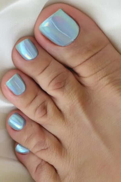 Zbliżenie na pedicure z chromowanym efektem w pastelowym, niebiesko-perłowym odcieniu. Stylizacja przywodzi na myśl połysk muszli lub odbicie światła na wodzie. Paznokcie są równo wypiłowane i intensywnie błyszczące, idealnie oddające estetykę trendu „syrenich paznokci”.