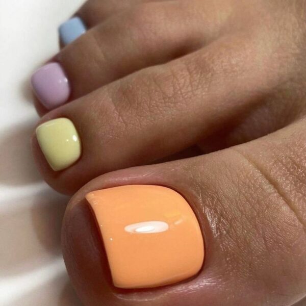 Pedicure na sierpień 2025: Pedicure w stylu pastel rainbow – każdy paznokieć pomalowany jest na inny pastelowy kolor: brzoskwiniowy, żółty, fioletowy i błękitny. Stylizacja przywodzi na myśl sorbetowe odcienie i wakacyjną beztroskę. Paznokcie są błyszczące, krótkie i zadbane, z równym kształtem płytek.