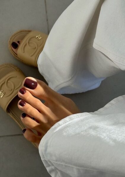 Pedicure na jesień 2025: paznokcie u stóp w intensywnym, ciemnoczerwonym odcieniu, zestawione z jasnymi spodniami i beżowymi klapkami Chanel. Stylizacja łącząca luksusowy charakter z ponadczasową elegancją.