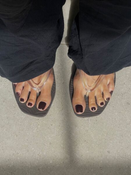 Klasyczny brązowy pedicure – głębokie, ciemne paznokcie u stóp z połyskiem, widoczne w transparentnych klapkach. Stonowana stylizacja w czerni podkreśla wyrazisty kolor lakieru.