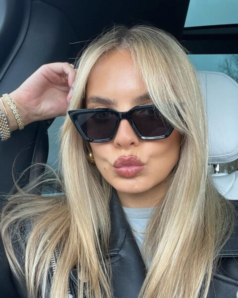 Zdjęcie kobiety siedzącej w sachodzie. Robi dziubek ustami. Ma długie, blond włosy rozpuszczone na ramiona. Ma na nosie okulary przeciwsłoneczne. Ubrana jest w szarą bluzkę i skórzaną, czarną kurtkę. Unosi rękę do głowy i ma na ręce dużo złotych bransoletek.