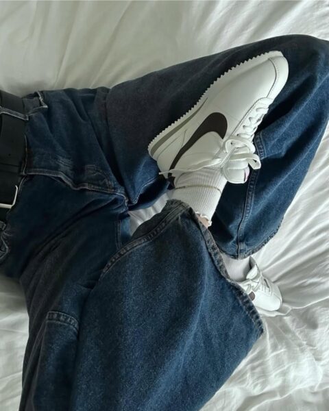 Jak rozpoznać oryginalne sneakersy? Na zdjęciu widać kobietę leżącą na łóżku w białej pościeli. Widać jej nogi ubrane w jeansy i białe sneakersy Nike.