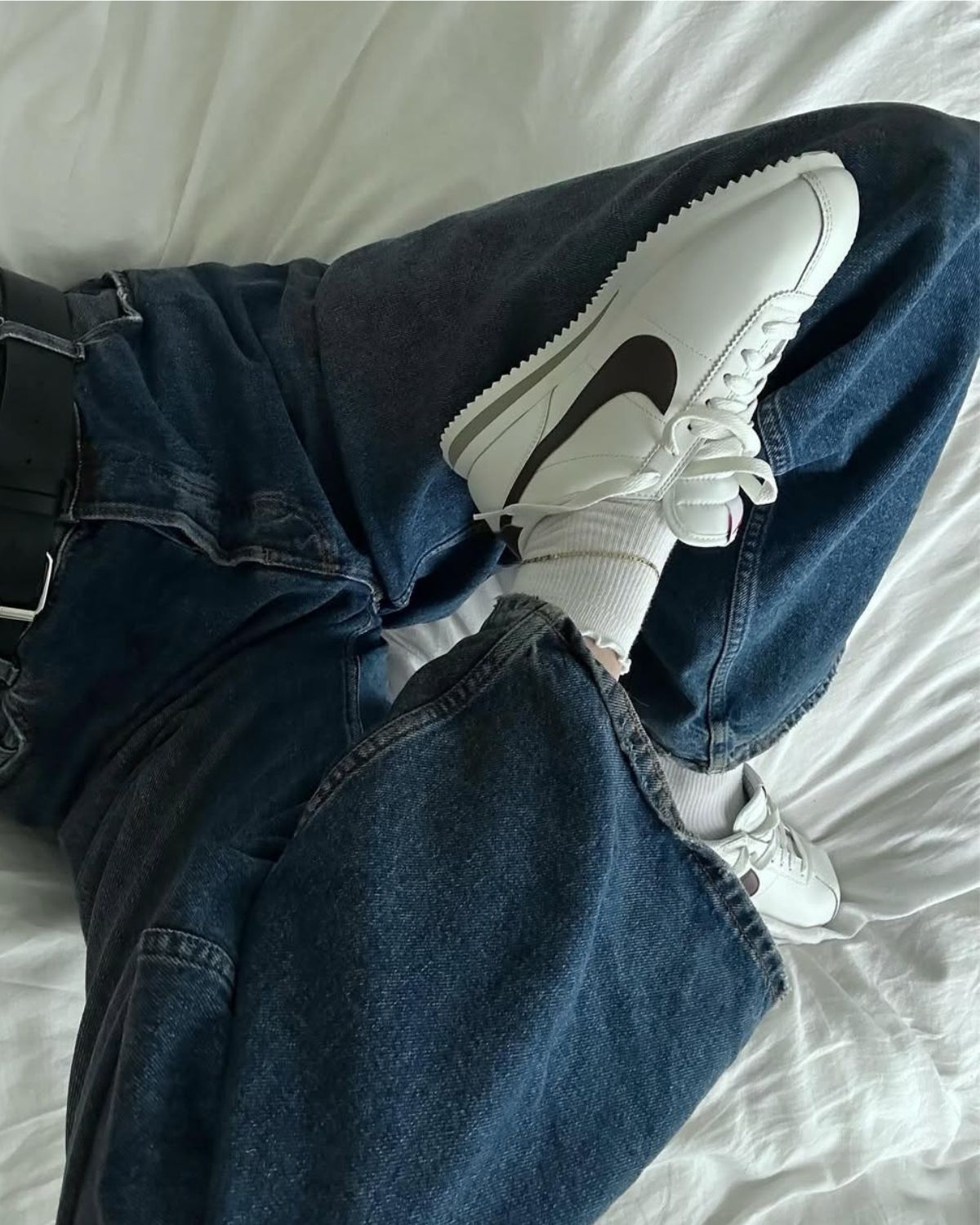 Jak rozpoznać oryginalne sneakersy? Na zdjęciu widać kobietę leżącą na łóżku w białej pościeli. Widać jej nogi ubrane w jeansy i białe sneakersy Nike.