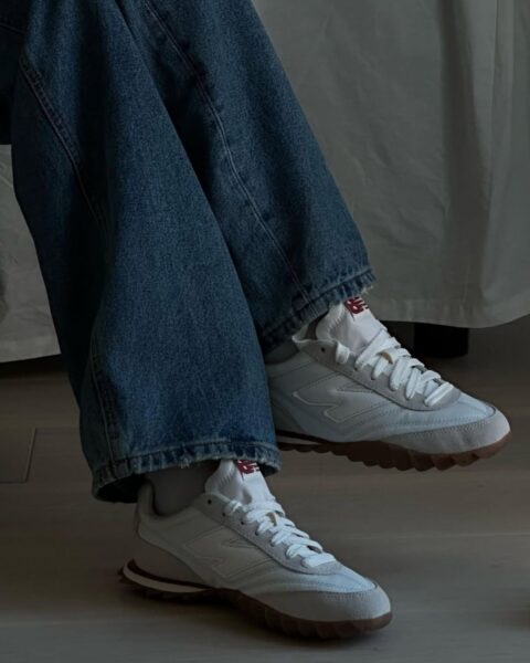 Zdjęcie ze zbliżeniem na białe, oryginalne sneakersy marki New Balance. Widać nogawki niebieskich jeansów i nogę założoną na nogę. 