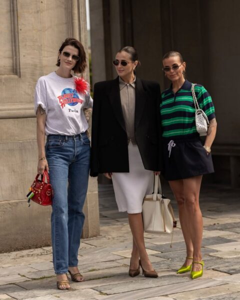 Zdjęcie kobiet na fashion week w Kopenhadze. Pokazują, jak zacząć przygodę z modą, czyli inwestując w modną garderobę. Pierwsza od lewej ma krótkie włosy, biały T-shirt z nadrukiem i czerwoną, kwiatową broszkę, jeansy, sandałki na obcasie oraz czerwoną torebkę. W środku jest brunetka z kokiem, okularami przeciwsłonecznymi, ubrana w beżową koszulę, białą ołówkową spódnicę, czarną marynarkę oversize i dużą, kremową torbę. Trzecia kobieta również ma gładko zaczesane w kok włosy, okulary przeciwsłoneczne, koszulkę polo w zielono-granatowe paski, czarne szorty, zielone czółenka oraz białą torebkę w groszki.