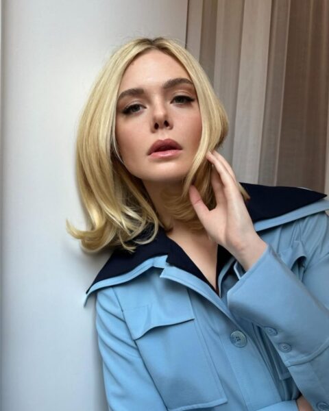 Zdjęcie aktorki Elle Fanning, która ma na głowie modny kolor lemon platinum blonde, czyli cytrynowy, platynowy blond. Ubrana jest w niebieską koszulę z szerokim, granatowym kołnierzem. Ma włosy do ramion, lekko podwinięte. Stoi przy ścianie, przykłada rękę do twarzy.