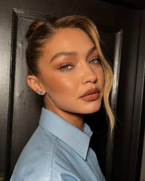 Zdjęcie modelki Gigi Hadid. Ma włosy spięte w koka i z grzywką side part, czyli na bok. Ubrana jest w niebieską, skórzaną kurtkę. Stoi bokiem, ale obraca twarz w stronę obiektywu.