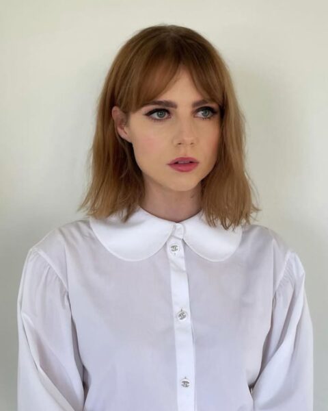 Zdjęcie Lucy Boynton. Ma ciemnoblond włosy do ramion i grzywkę curtain bangs. Ubrana jest w białą koszulę z kołnierzykiem z zaokrąglonymi rogami