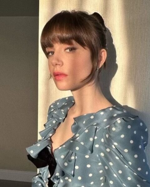 Zdjęcie aktorki Lily Collins. Ma włosy spięte w kok i grzywkę typu bunny bangs. Ubrana jest w niebieską bluzkę z falbankami i w groszki. 