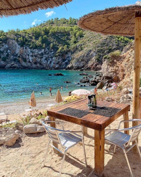 Zdjęcie brzegu morza na wyspie Ibiza. Przy brzegu znajduje się, widoczny na pierwszym planie, stół z krzesłami. Nad nim są parasole. W tle widać zatokę oraz nadmorskie skały.