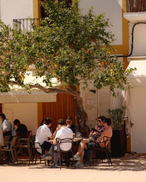Zdjęcie ogródka restauracyjnego przy placu na Ibizie. Widać grupę ludzi siedzących przy stoliku, nad nimi jest drzewo cytrynowe, obok zadaszenie restauracji.