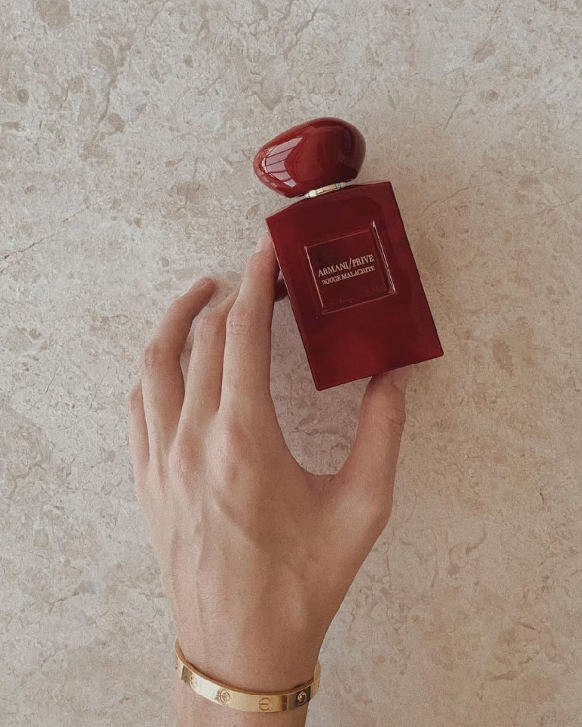 Zdjęcie przedstawiające najpiękniejsze perfumy na jesień 2025. Są w czerwonym flakonie. Leżą na beżowym, marmurowym blacie. Trzymane są w dłoni, która ma złotą bransoletkę Cartier na nadgarstku.