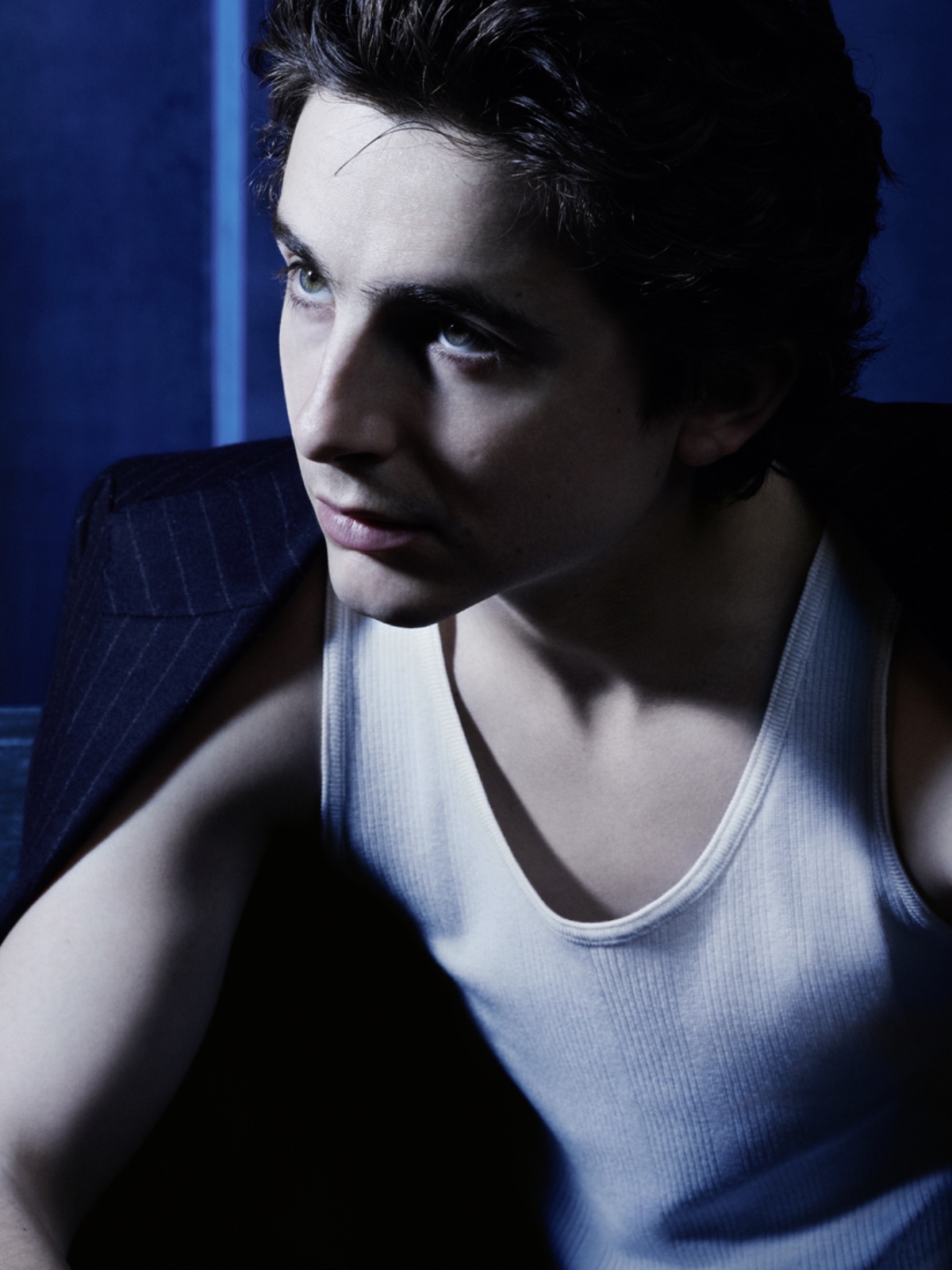 Portret Timothée Chalameta w kampanii Bleu de Chanel. Aktor w białej koszulce bez rękawów i granatowej marynarce narzuconej na ramiona patrzy w górę, w bok, podświetlony zimnym, niebieskim światłem. Jego spojrzenie podkreśla atmosferę intensywności i nieuchwytności.