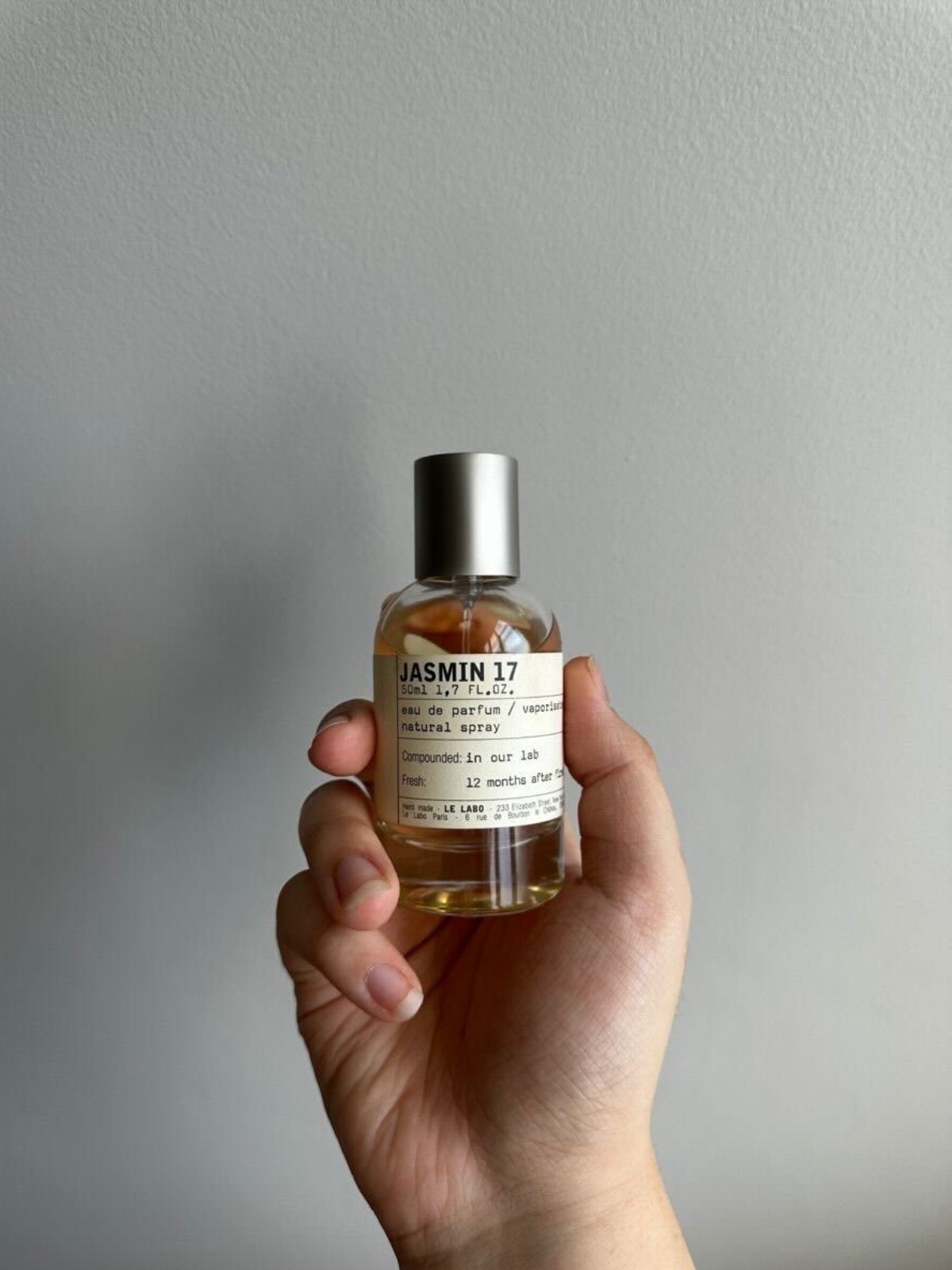 Najpiękniejsze jaśminowe perfumy: Butelka perfum Le Labo Jasmin 17 Eau de Parfum trzymana w dłoni – szklany, przezroczysty flakon w rozmiarze 50 ml, wypełniony złocistym płynem; srebrny korek i prosta etykieta w stylu laboratoryjnym kontrastują z ciepłym odcieniem zapachu.