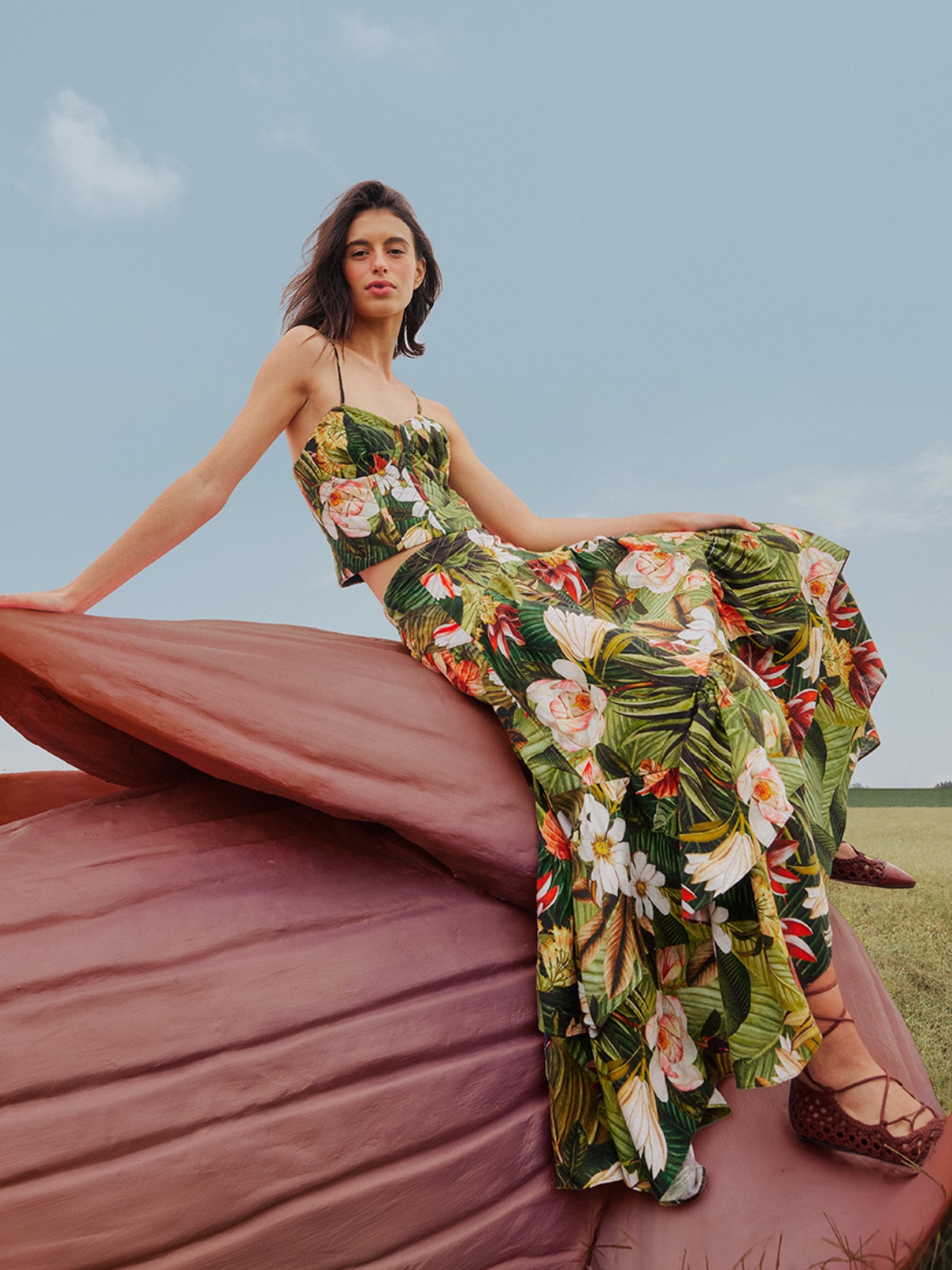 Sportofino trendy lato 2025: Modelka siedząca na rzeźbie w kształcie gigantycznego pąka kwiatu, ubrana w zestaw crop top i długą spódnicę w tropikalny print z liśćmi palm i dużymi, białymi kwiatami. Tło stanowi błękitne niebo, co podkreśla letni klimat stylizacji.